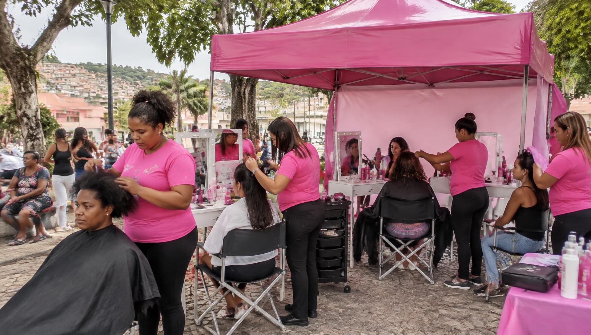 Ação de beleza comunitária em praça, com tenda rosa, estações com espelhos e equipe uniformizada atendendo mulheres